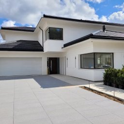 Onbud sp z o.o. - Nowoczesny dom jednorodzinny z białą elewacją, czarnym dachem i dużym garażem, otoczony kostką brukową i świeżo zasadzonymi krzewami w donicach.