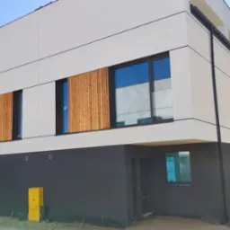 Nowoczesny dom z białą elewacją i ciemnoszarym cokołem, drewniane panele dekoracyjne na oknach, błękitne niebo w tle.