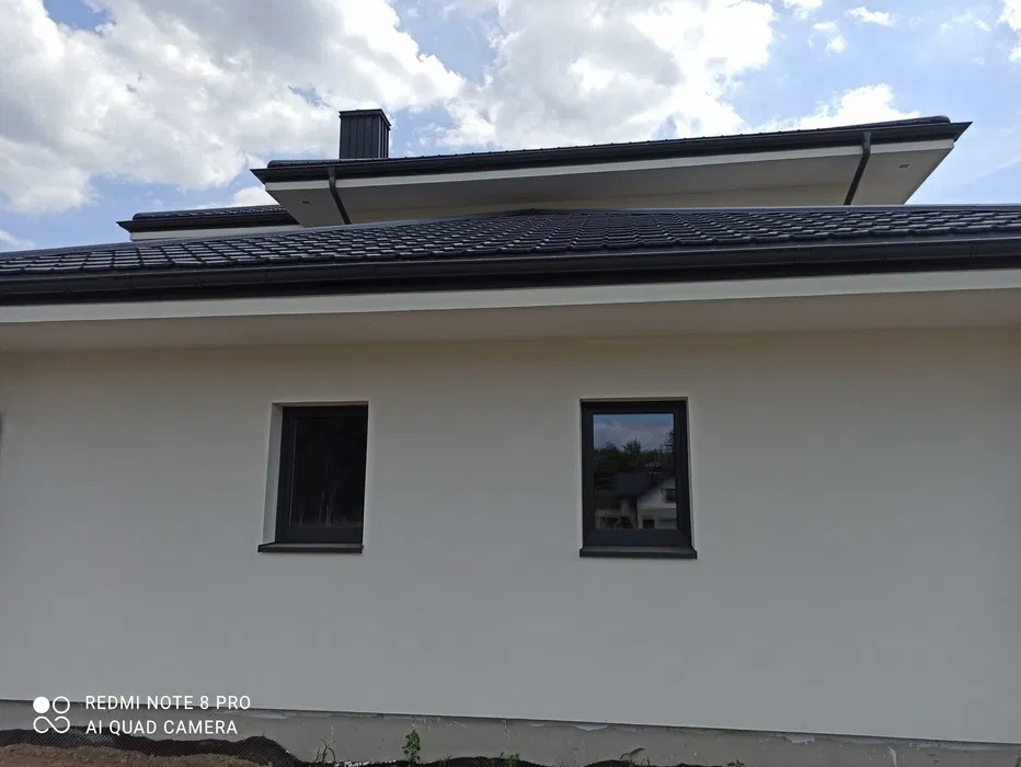 Świeżo otynkowana elewacja domu jednorodzinnego z dwoma oknami w ciemnych ramach, widok z boku, z widocznym czarnym dachem i kominem na tle błękitnego nieba z chmurami.