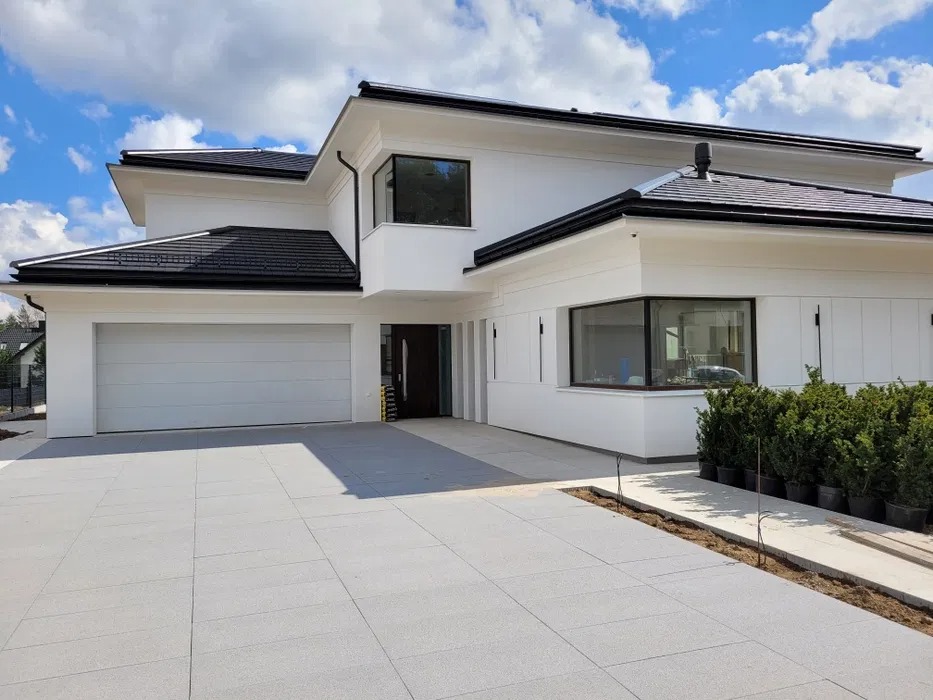 Nowoczesny dom jednorodzinny z białą elewacją, czarnym dachem i dużym garażem, otoczony kostką brukową i świeżo zasadzonymi krzewami w donicach.