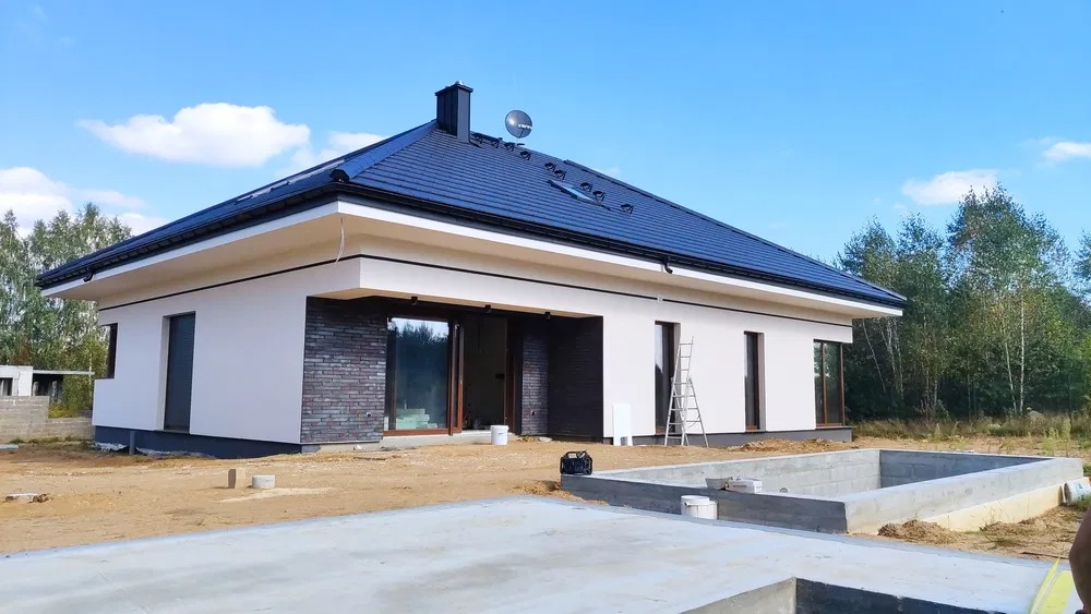 Nowoczesny dom jednorodzinny z białą elewacją, fragmentem ściany wykończonym cegłą, ciemnym dachem i satelitą, w trakcie budowy, z widocznym fundamentem basenu.
