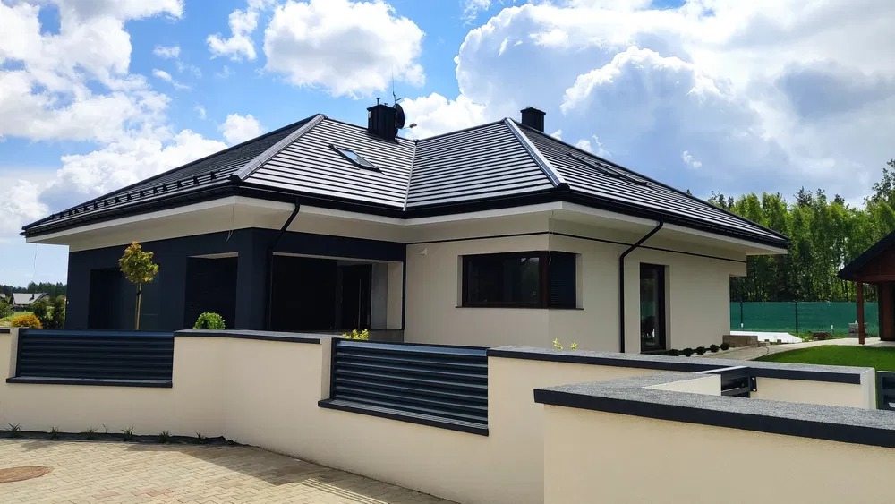 Nowoczesny dom jednorodzinny z czarnym dachem i elementami elewacji, otoczony niskim murem z metalowym ogrodzeniem, na tle błękitnego nieba z obłokami.