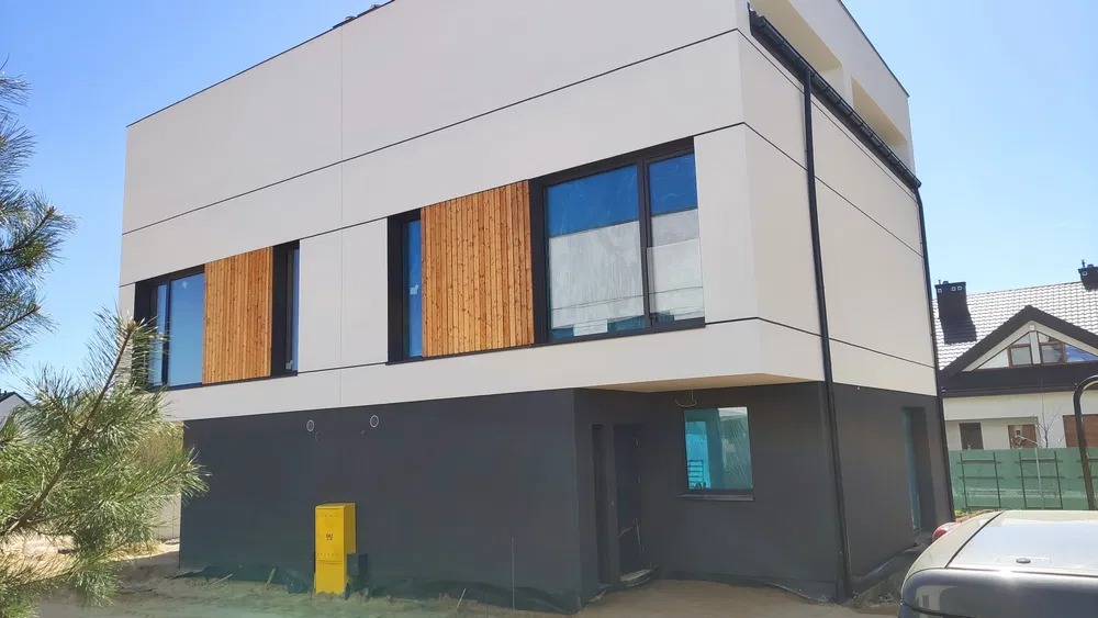 Nowoczesny dom z białą elewacją i ciemnoszarym cokołem, drewniane panele dekoracyjne na oknach, błękitne niebo w tle.