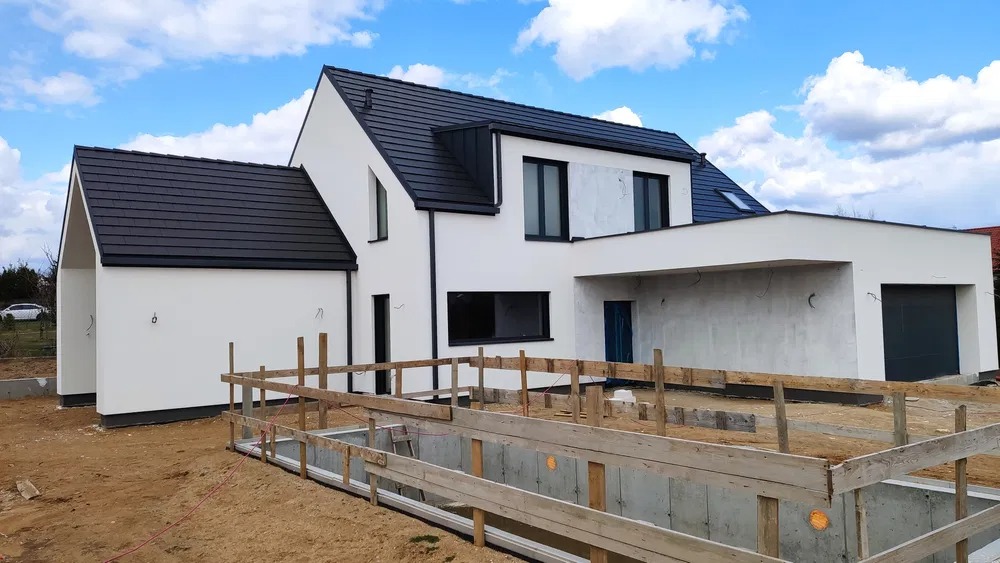 Nowoczesny dom jednorodzinny z czarnym dachem i białą elewacją w trakcie budowy, widoczne deskowanie fundamentów i częściowo wykończona elewacja.