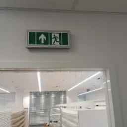 Oznakowanie drogi ewakuacyjnej z zielonym piktogramem na białej ścianie nad drzwiami w nowym sklepie z regałami w tle.