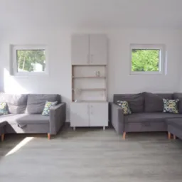 Jasne wnętrze z dwoma szarymi narożnikami, białą szafką i dwoma oknami z widokiem na zieleń.