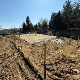 Usługi Geodezyjne Geo-Progres Sławomir Wolanin - Wytyczony teren pod budowę domu jednorodzinnego z widocznymi drewnianymi słupkami i niwelacją terenu, w tle drzewa i zabudowania.