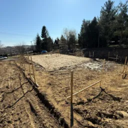 Wytyczony teren pod budowę domu jednorodzinnego z widocznymi drewnianymi słupkami i niwelacją terenu, w tle drzewa i zabudowania.