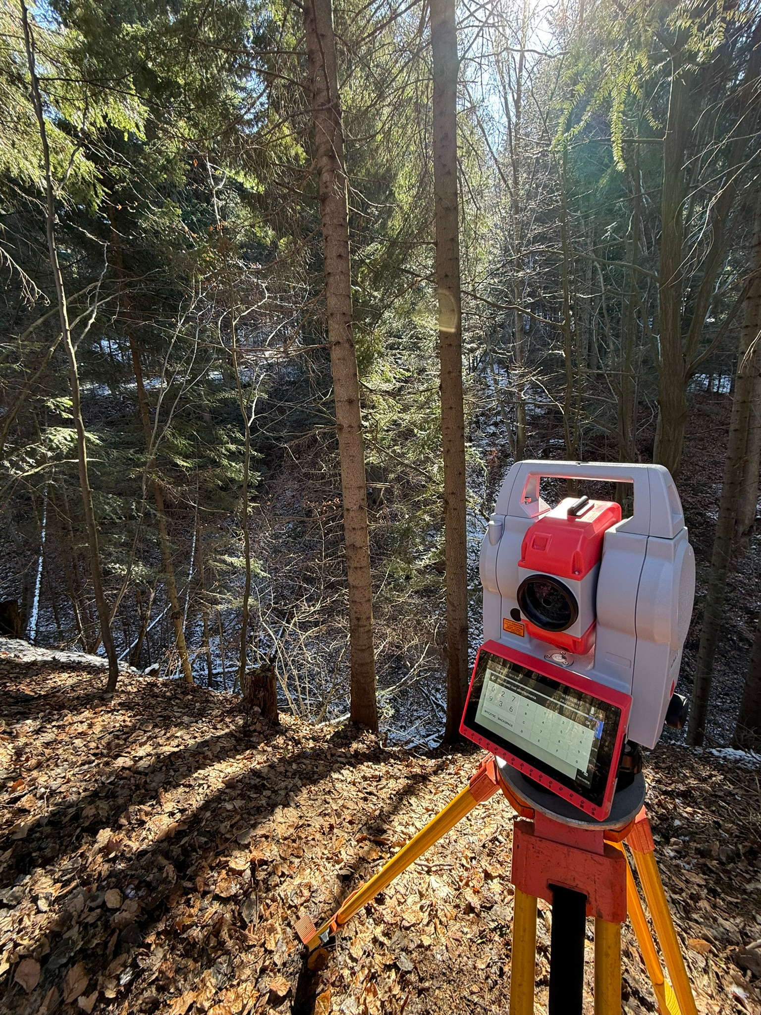 Teodolit elektroniczny ustawiony na trójnogu w lesie, widoczny ekran dotykowy z interfejsem oprogramowania geodezyjnego, w tle drzewa i opadłe liście.