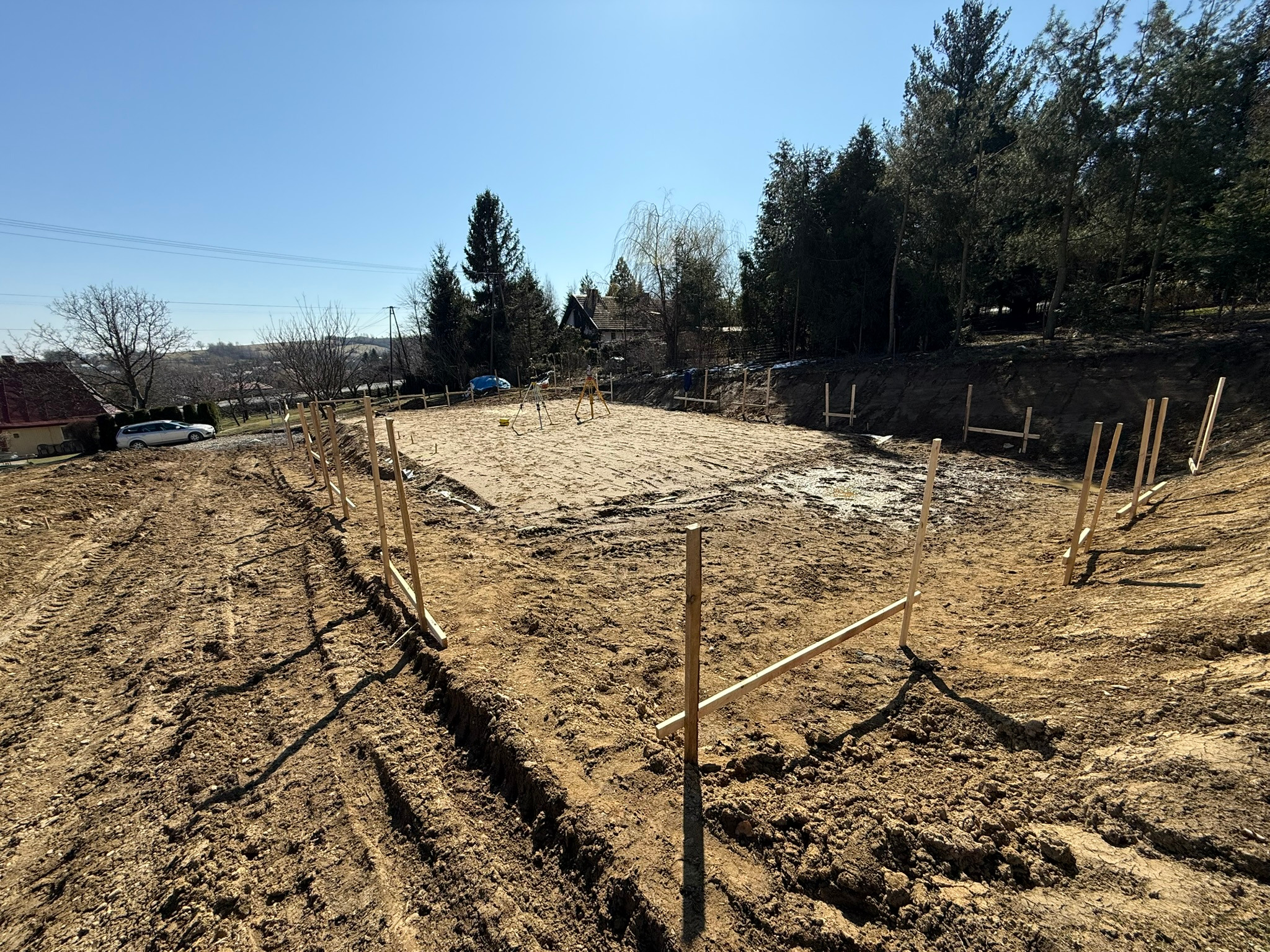 Wytyczony teren pod budowę domu jednorodzinnego z widocznymi drewnianymi słupkami i niwelacją terenu, w tle drzewa i zabudowania.