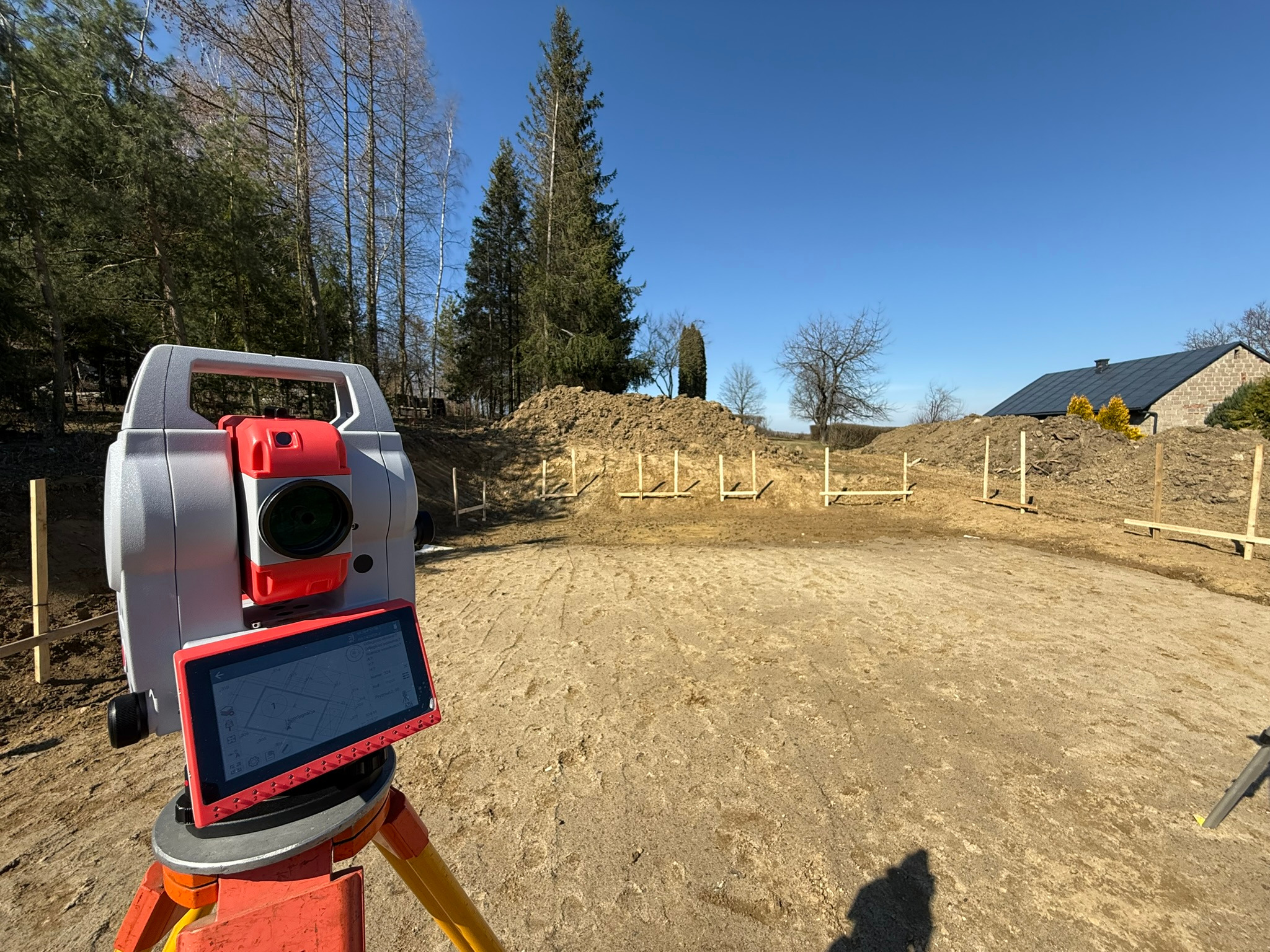 Profesjonalny niwelator elektroniczny ustawiony na placu budowy z widocznym ekranem dotykowym wyświetlającym plan działki, w tle wytyczony teren pod fundamenty i drzewa.