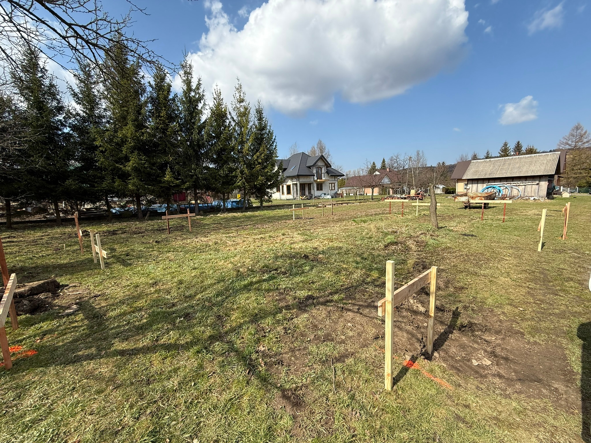 Wytyczanie fundamentów pod budowę domu jednorodzinnego, widoczne drewniane słupki z pomarańczowymi oznaczeniami na trawiastym terenie działki w słoneczny dzień.