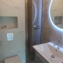 Nowoczesna łazienka z umywalką na szafce, podświetlanym lustrem w kształcie okręgu, zabudowaną szafką i toaletą w jasnych odcieniach szarości i bieli z marmurkowym wzorem na płytkach.