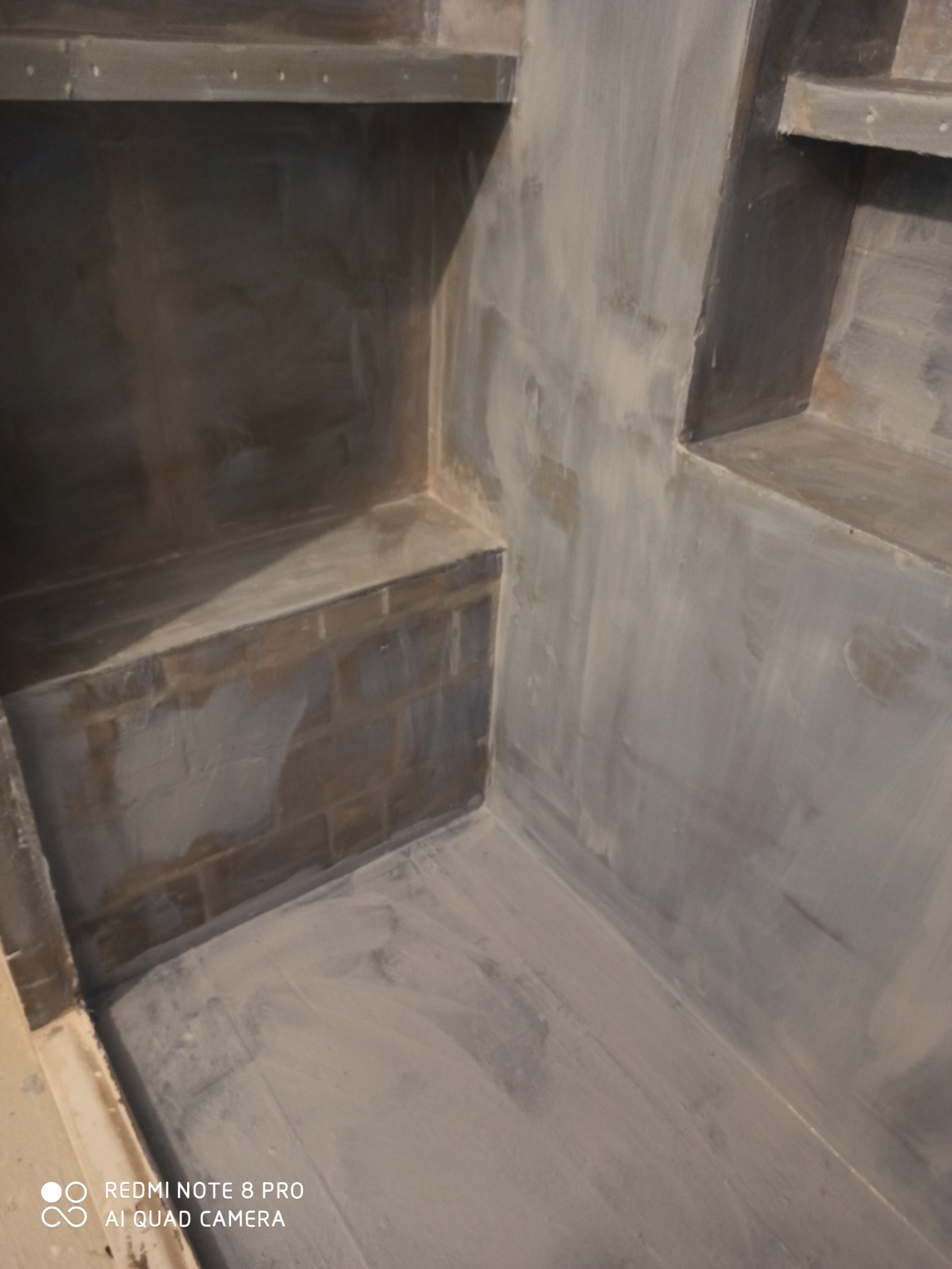 Wnętrze wnęki z półkami w trakcie przygotowania do dalszych prac wykończeniowych, ściany i podłoga pokryte warstwą szarego podkładu.