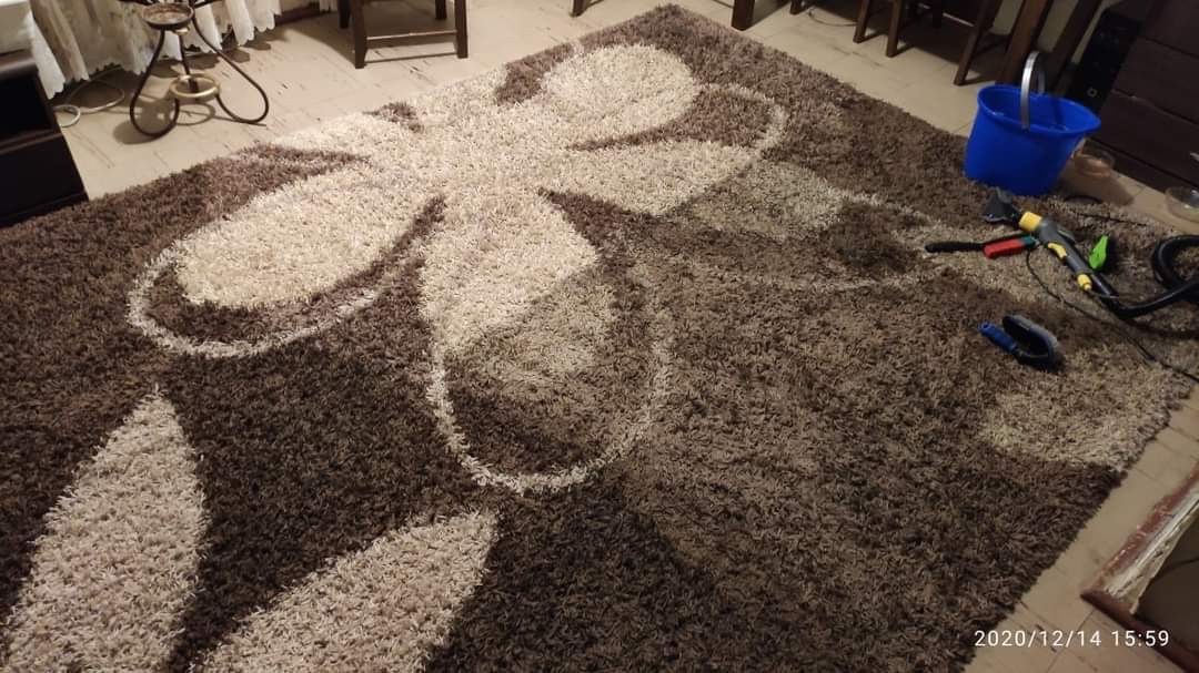 Wielki, brązowo-beżowy dywan shaggy z motywem kwiatowym, na którym leżą narzędzia do czyszczenia: szczotka, spryskiwacz i niebieskie wiadro z wodą.