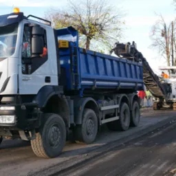 Ciężarówka Iveco z niebieską skrzynią ładunkową podczas frezowania asfaltu na ulicy, w tle widoczna frezarka i pracownicy w odblaskowych ubraniach.
