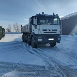 Ciężarówka Iveco Trakker na śniegu z widocznymi śladami opon, zielone kontenery i budynek w tle, słoneczny dzień.