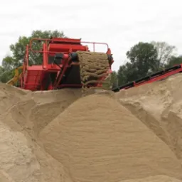 Czerwona przesiewarka do kruszywa wysypuje materiał na dużą pryzmę. Widoczne taśmociągi i zieleń w tle.