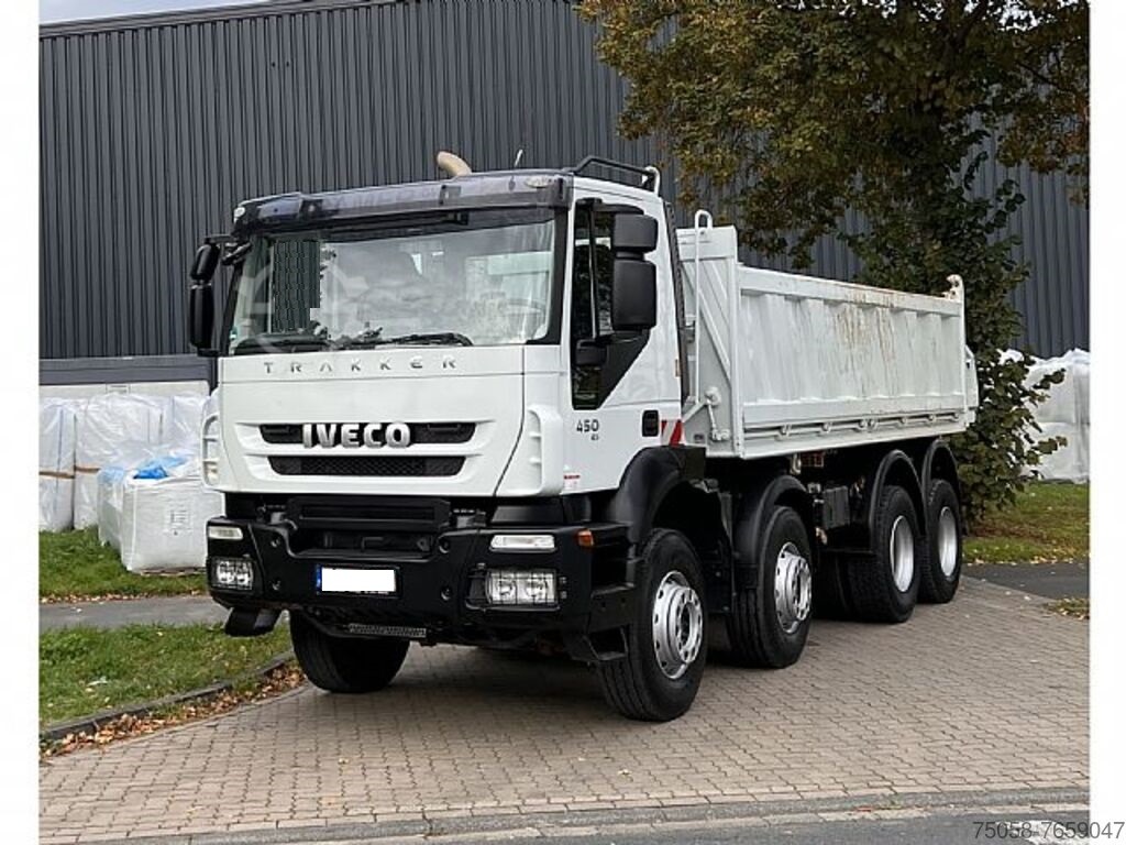 Biała ciężarówka wywrotka Iveco Trakker 450, zaparkowana na kostce brukowej, z widocznymi śladami użytkowania na skrzyni ładunkowej, na tle szarej hali i drzewa.