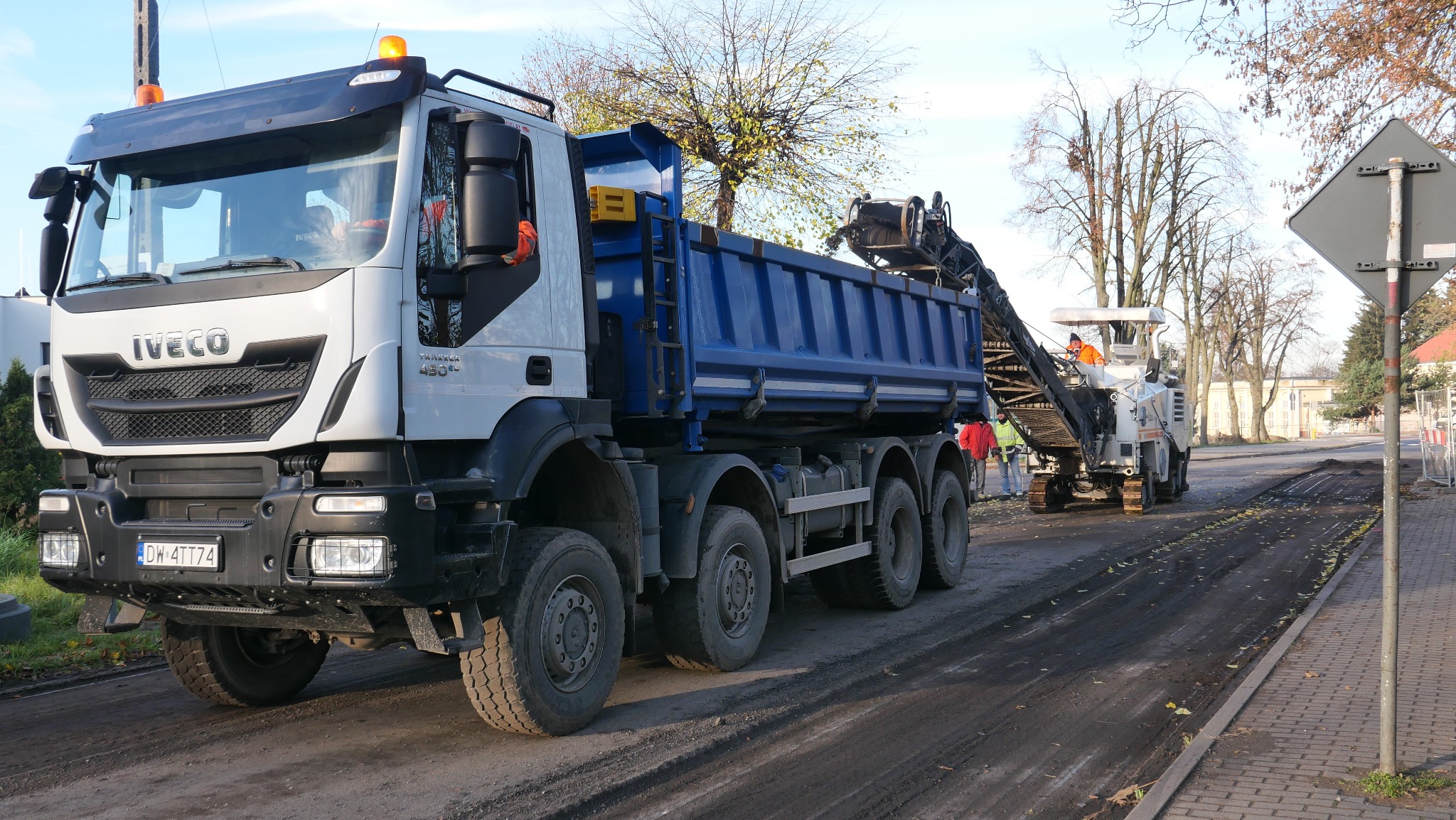 Ciężarówka Iveco z niebieską skrzynią ładunkową podczas frezowania asfaltu na ulicy, w tle widoczna frezarka i pracownicy w odblaskowych ubraniach.