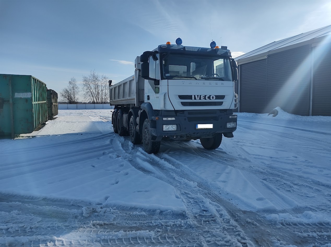 Ciężarówka Iveco Trakker na śniegu z widocznymi śladami opon, zielone kontenery i budynek w tle, słoneczny dzień.