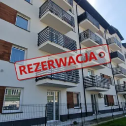 Nowoczesny budynek apartamentowy z balkonami i elementami dekoracyjnymi z drewna, na elewacji widnieje czerwony napis 'REZERWACJA', w tle parking i zieleń.