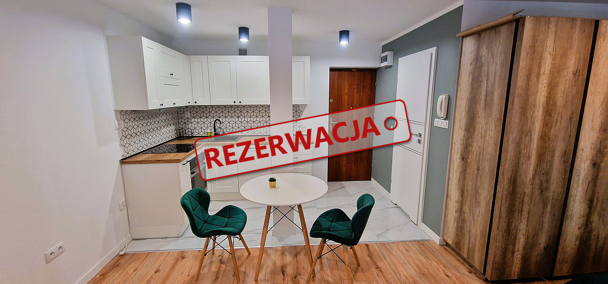 Wnętrze mieszkania z aneksem kuchennym i okrągłym stołem, na zdjęciu widoczny napis 'REZERWACJA'.