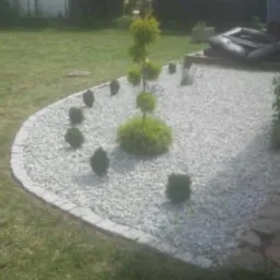 Ogród z białym żwirem, obrzeżem z kostki i formowanymi krzewami na trawniku przed domem.