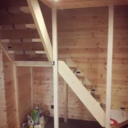Wnętrze domu w Leśniewie w trakcie budowy, widoczna drewniana konstrukcja schodów i ścian, elementy izolacji oraz materiały budowlane w pomieszczeniu pod schodami.