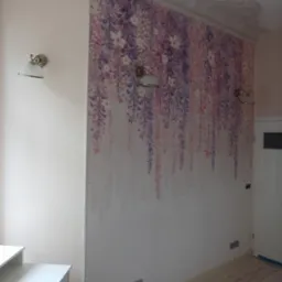 Ściana w pokoju z tapetą imitującą kwiaty glicynii, oświetlona dwoma kinkietami w stylu retro, z widoczną dekoracyjną sztukaterią sufitową.