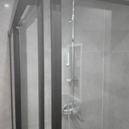 Sp.zo.o LVASKO - Nowoczesna kabina prysznicowa z czarnymi profilami i szklanymi drzwiami, widoczna bateria i deszczownica na tle szarych płytek.