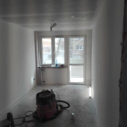 Sp.zo.o LVASKO - Pokój w trakcie odświeżania: zagruntowane ściany, surowy sufit, widoczny odkurzacz przemysłowy, drabina oraz prowizoryczne oświetlenie halogenowe pod oknem.