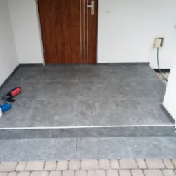 Wyłożony szarymi płytkami gresowymi taras przed wejściem do domu, widoczne narzędzia pracy i fragment kostki brukowej.