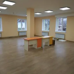 Jasne, przestronne wnętrze biurowe z drewnianą podłogą, pomalowanymi na beżowo ścianami, białymi grzejnikami i oknami oraz dwoma małymi biurkami w kolorach pomarańczowym i żółtym.