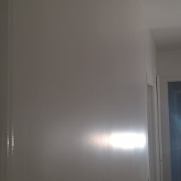 Sp.zo.o LVASKO - Świeżo pomalowana, gładka, biała ściana z widocznym połyskiem farby, oświetlona punktowo, z fragmentem białych drzwi po prawej stronie.