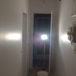 Sp.zo.o LVASKO - Korytarz w trakcie malowania na biało, widoczny reflektor budowlany na trójnogu, dwa wiadra z farbą i zabezpieczona folią podłoga.