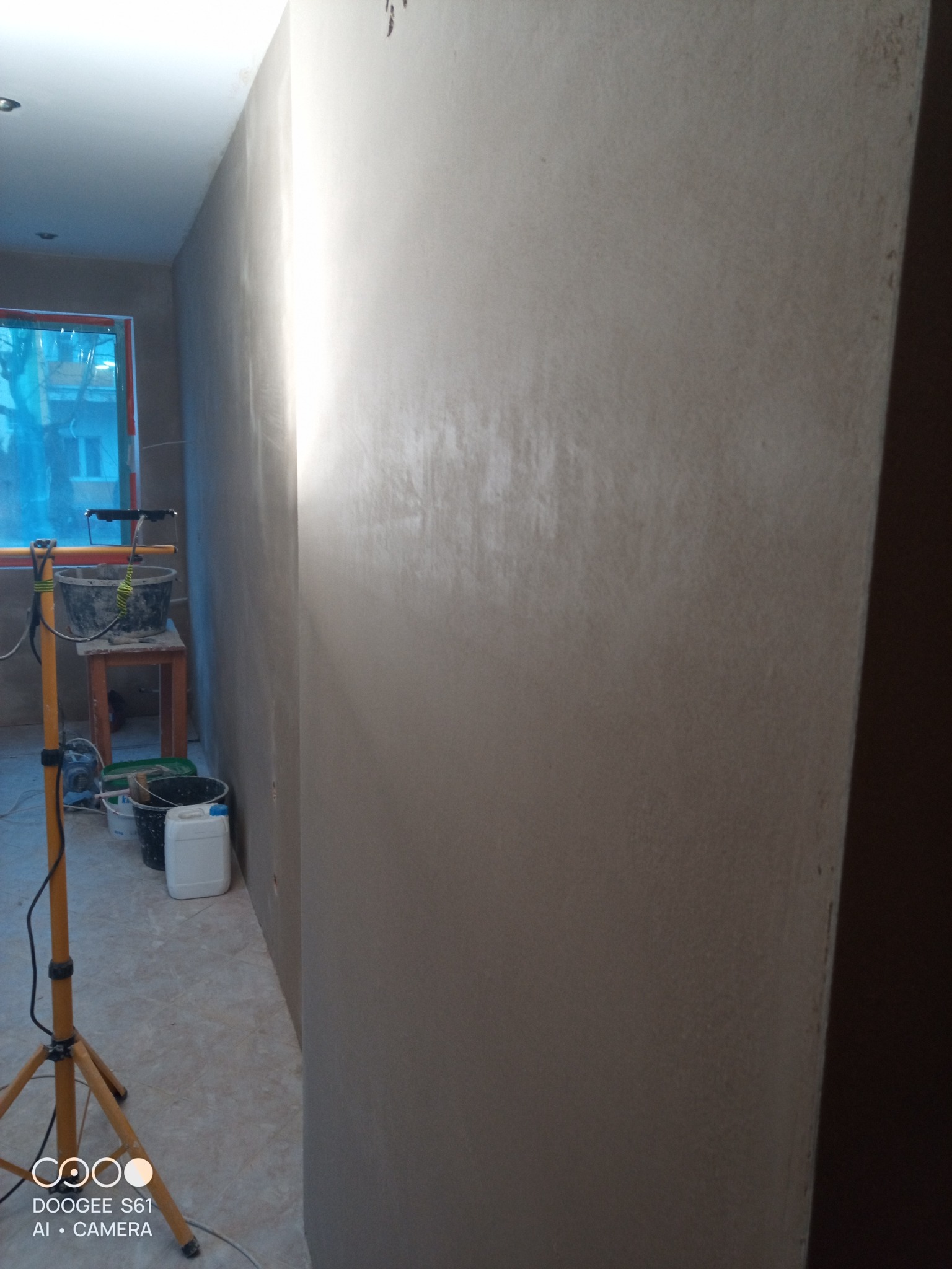 Ściana po nałożeniu gładzi, widoczne wiaderka, szpachelki i lampa na statywie w pomieszczeniu w trakcie prac wykończeniowych.