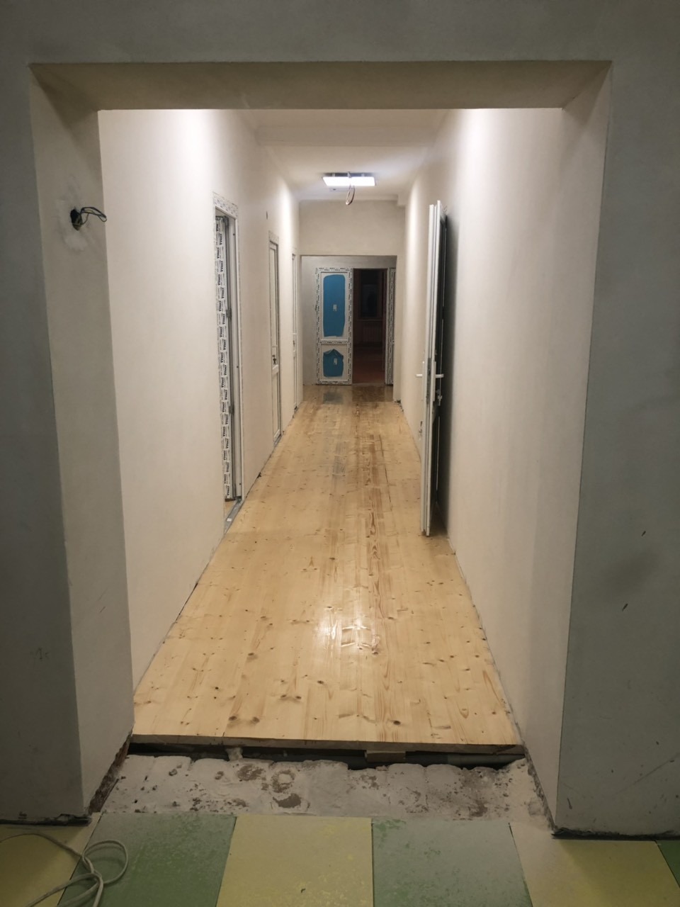 Korytarz w trakcie remontu: surowe drewniane deski podłogowe częściowo ułożone, ściany pomalowane na jasny kolor, widoczne przygotowania do dalszych prac wykończeniowych, kabel na podłodze.