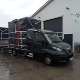 Czarny bus Iveco z platformą transportową przewozi dwa duże ładunki zabezpieczone czarną plandeką i czerwonymi pasami, na mokrym betonowym placu przed budynkiem z szarą bramą.