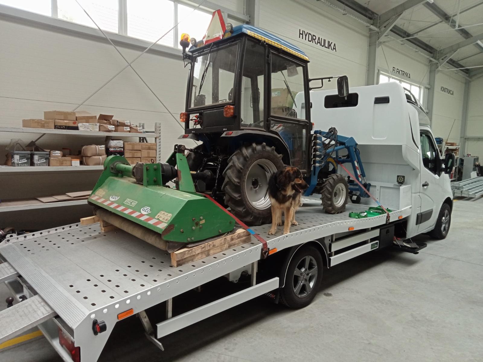 Ciągnik rolniczy z kosiarką na platformie pojazdu, zabezpieczony pasami, z psem stojącym obok, w hali z napisem HYDRAULIKA na ścianie.