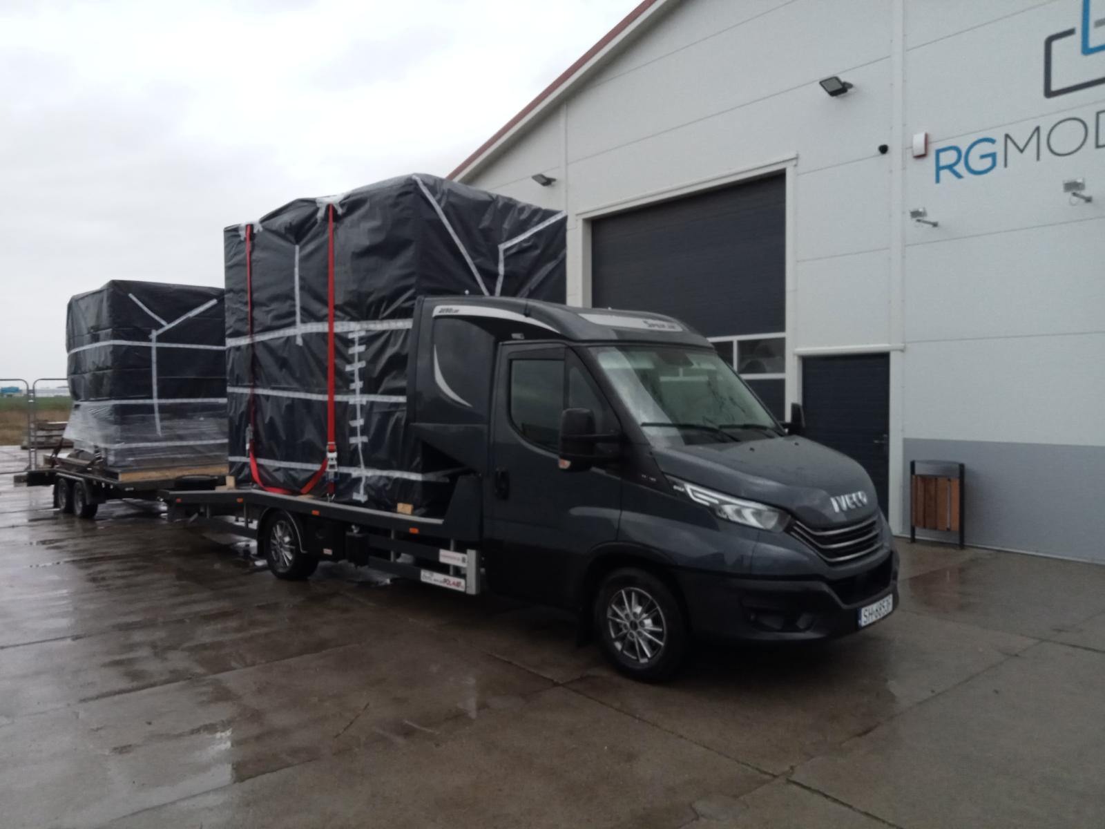 Czarny bus Iveco z platformą transportową przewozi dwa duże ładunki zabezpieczone czarną plandeką i czerwonymi pasami, na mokrym betonowym placu przed budynkiem z szarą bramą.