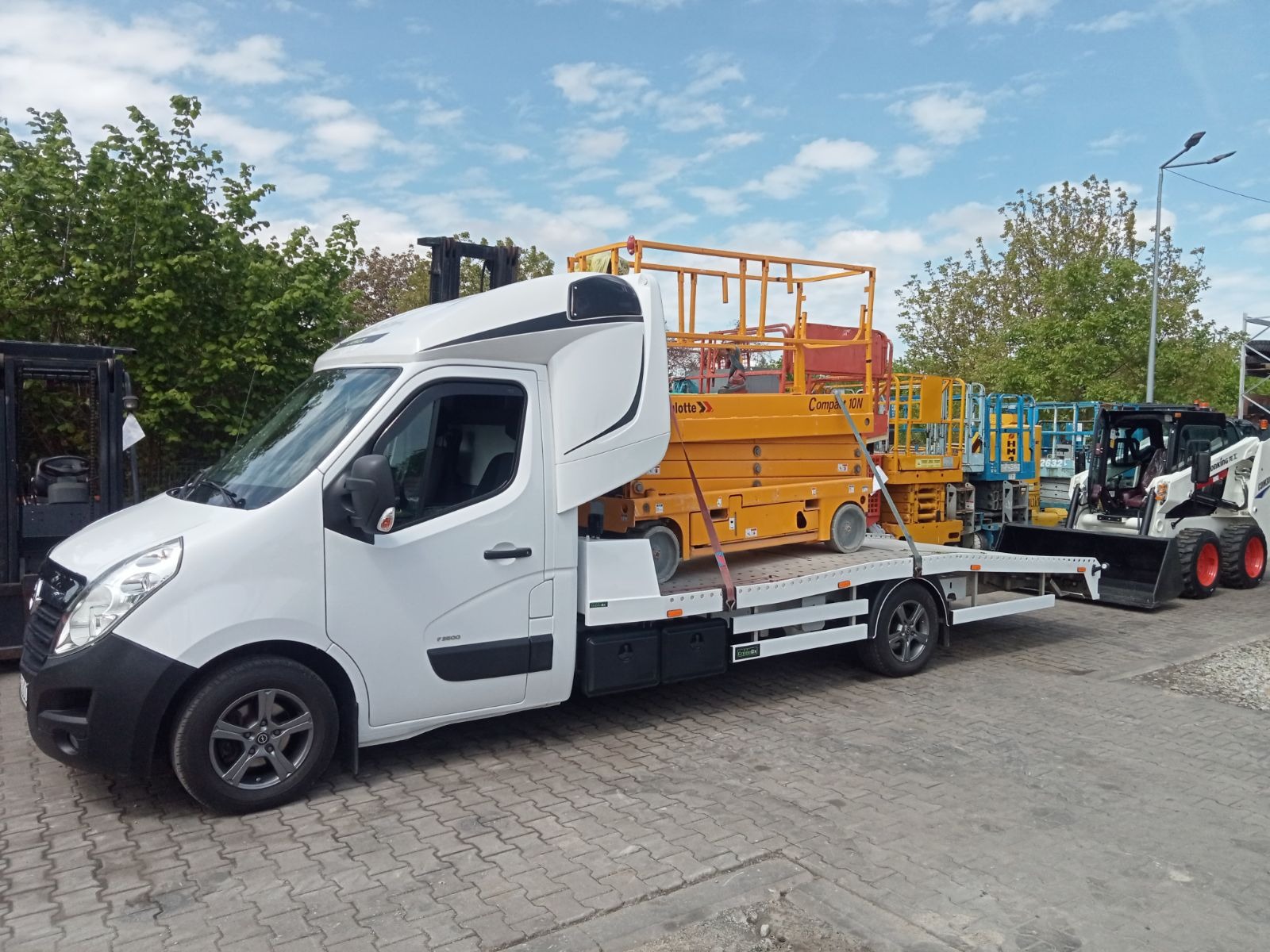 Biała laweta z platformą przewozi podnośnik nożycowy w kolorze żółtym oraz miniładowarkę Bobcat. Na platformie widoczne również inne podnośniki. W tle drzewa i częściowo widoczny wózek widłowy.