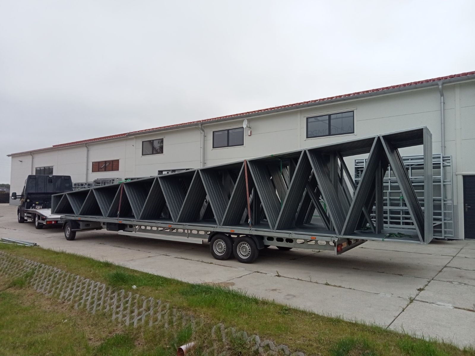 Czarny samochód z platformą transportuje długą, stalową konstrukcję kratownicową z trójkątnymi segmentami na tle białego budynku.