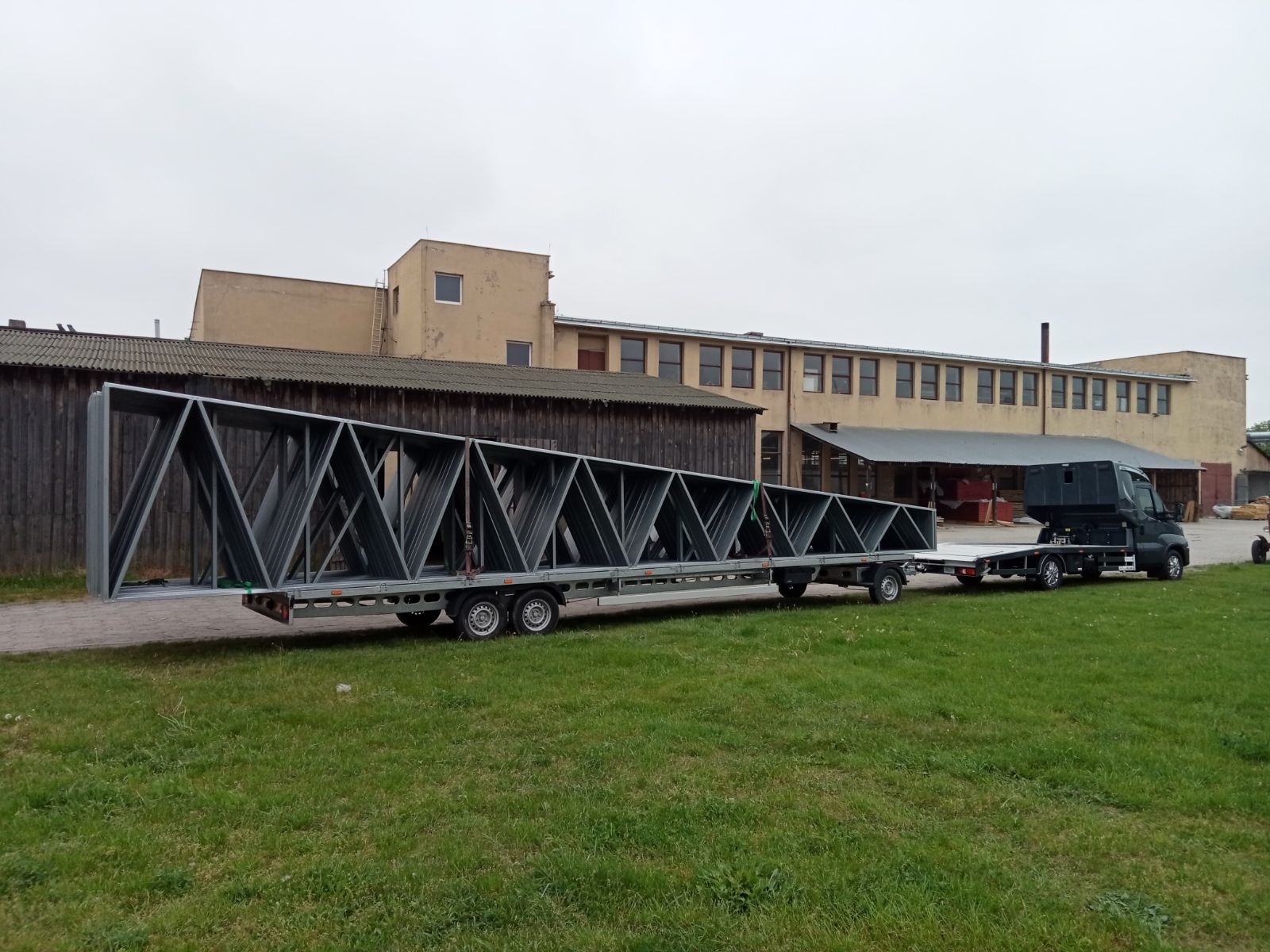 Czarny samochód dostawczy z naczepą przewozi długą, stalową konstrukcję kratownicową o trójkątnym wzorze na tle budynku przemysłowego z cegły.