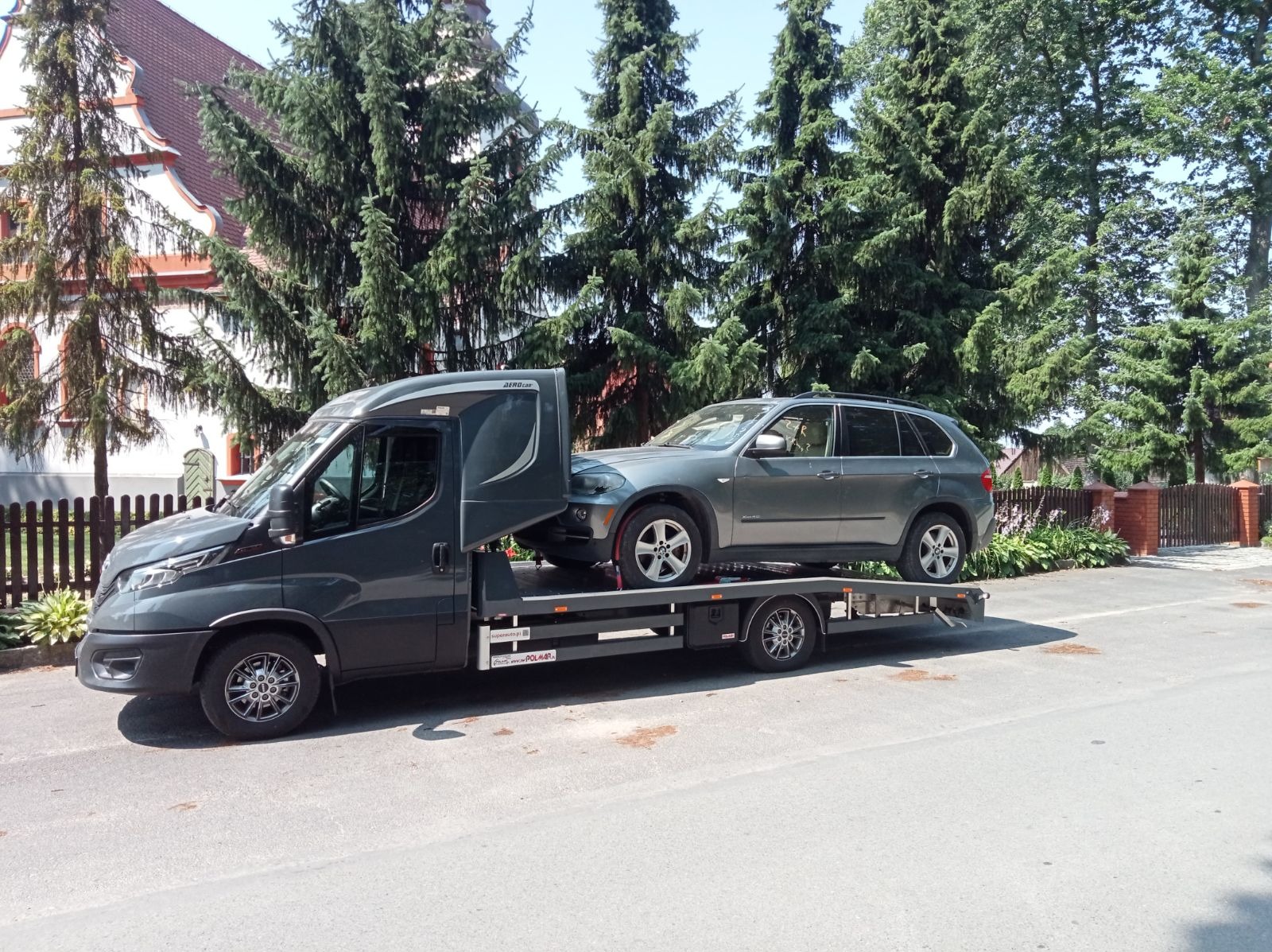 Szary samochód osobowy typu SUV przewożony na platformie lawety Iveco z zabudową Aerocar, na tle budynku z czerwonym dachem i zielonych drzew.