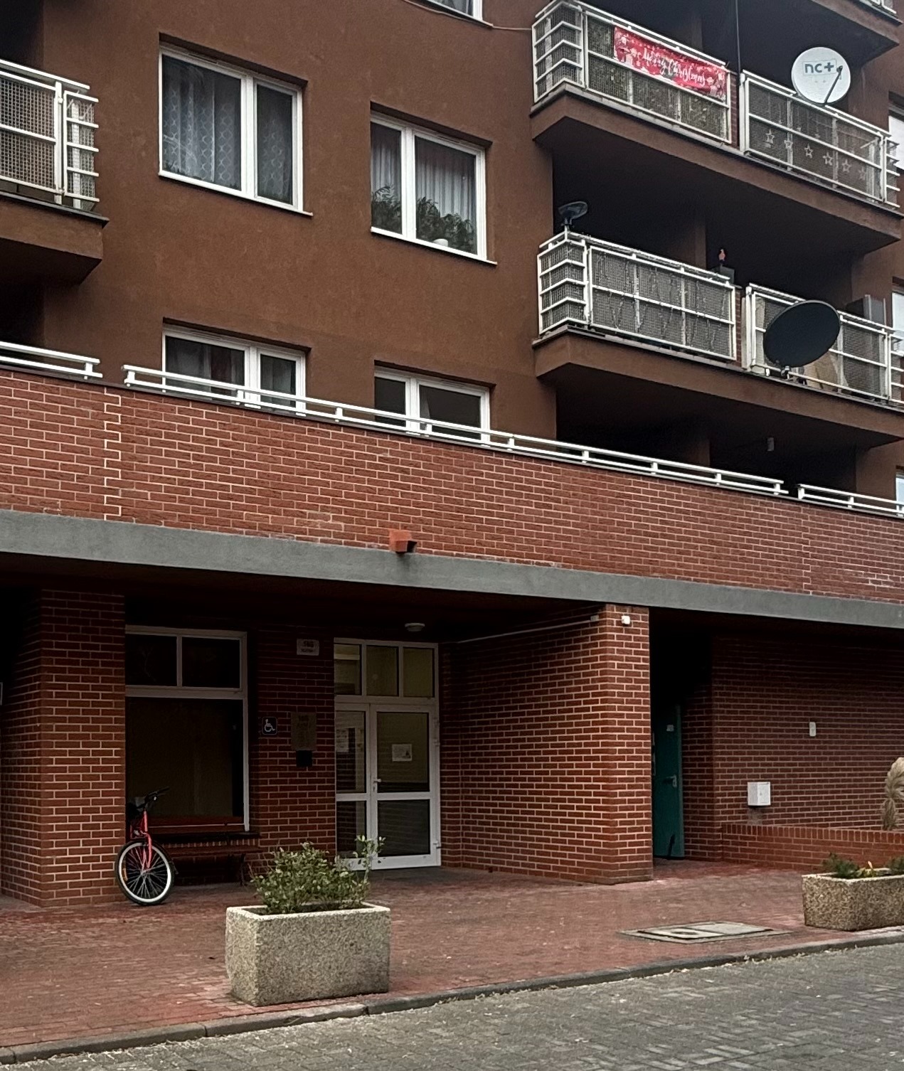 Wejście do budynku wielorodzinnego z czerwonej cegły, z rowerem opartym o ścianę i rośliną w kamiennej donicy. Balkony z antenami satelitarnymi na górnych piętrach.