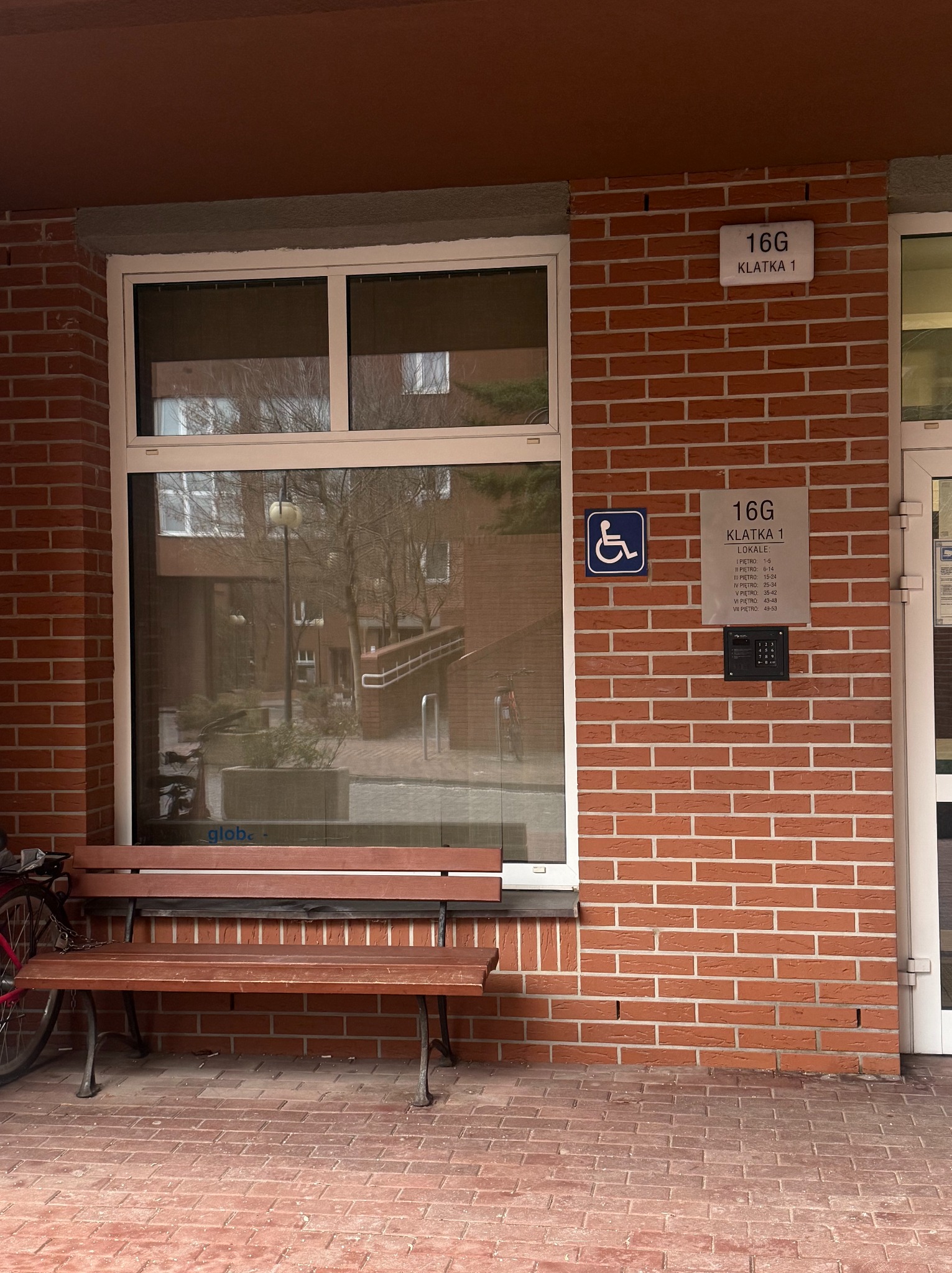Wejście do budynku z cegły z oznaczeniem '16G Klatka 1', ławką i symbolem dostępności dla osób niepełnosprawnych. Widoczne okno z odbiciem otoczenia.
