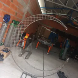 Półokrągła metalowa konstrukcja z siatki w warsztacie, oparta na metalowym wsporniku, butle z gazem w tle, surowa ceglana ściana.