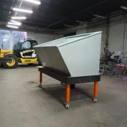 Szara metalowa skrzynia na konstrukcji z pomarańczowymi nogami na kółkach, w tle żółta ładowarka teleskopowa na hali.