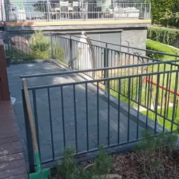 Metalowa balustrada pomalowana na szaro, zabezpieczająca taras z szarą kostką brukową, z widoczną łopatą opartą o balustradę i roślinnością w tle.